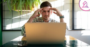 Hombre concentrado frente a una laptop, con las manos en la cabeza, trabajando en un entorno moderno con plantas y luz natural.
